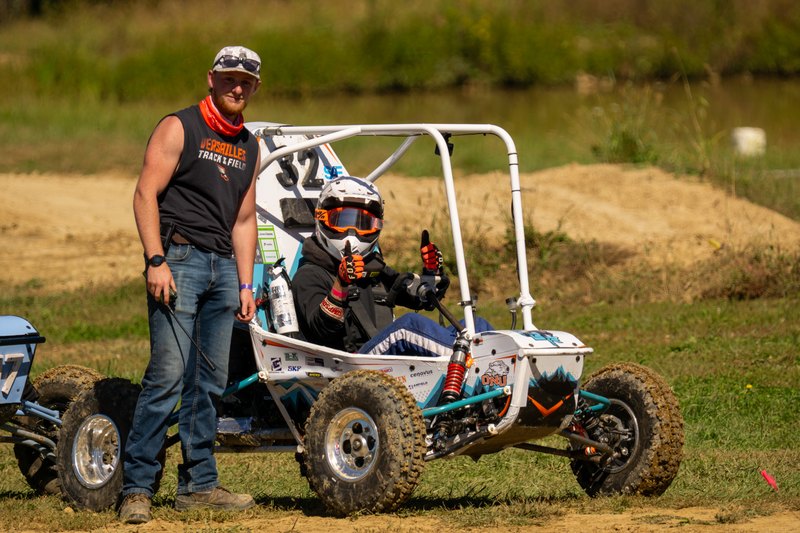 Suited-up driver in Baja car number 32 with a crew member standing beside it