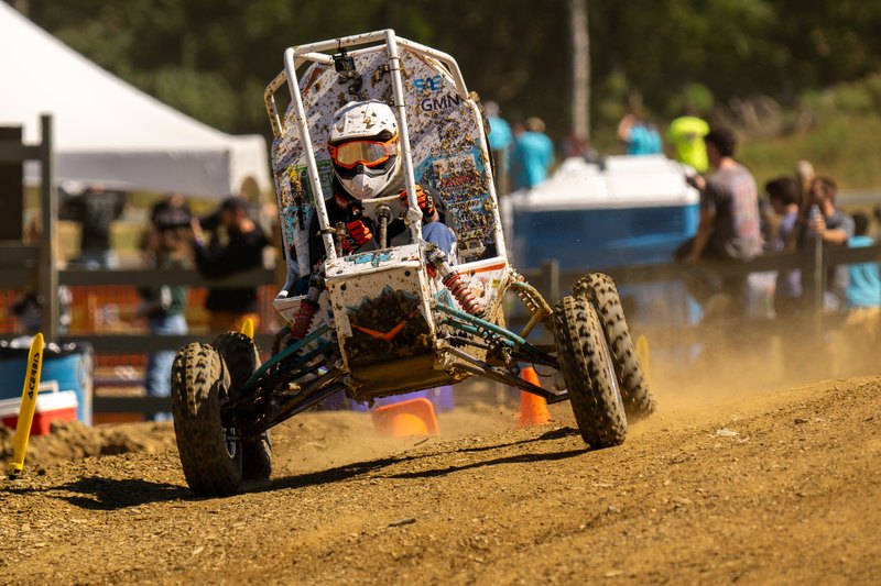 Baja car number 32 head-on over rough dirt with spectators and tents behind