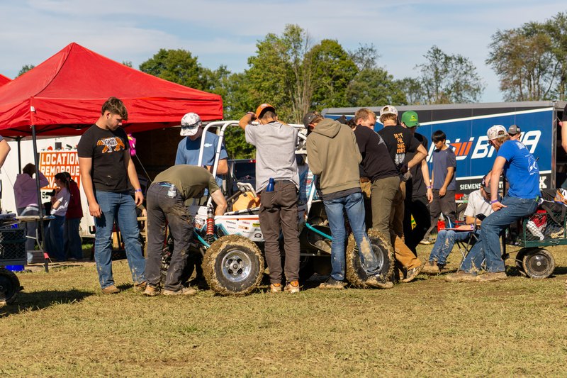 Team gathered around the Baja car at the pit under a red tent