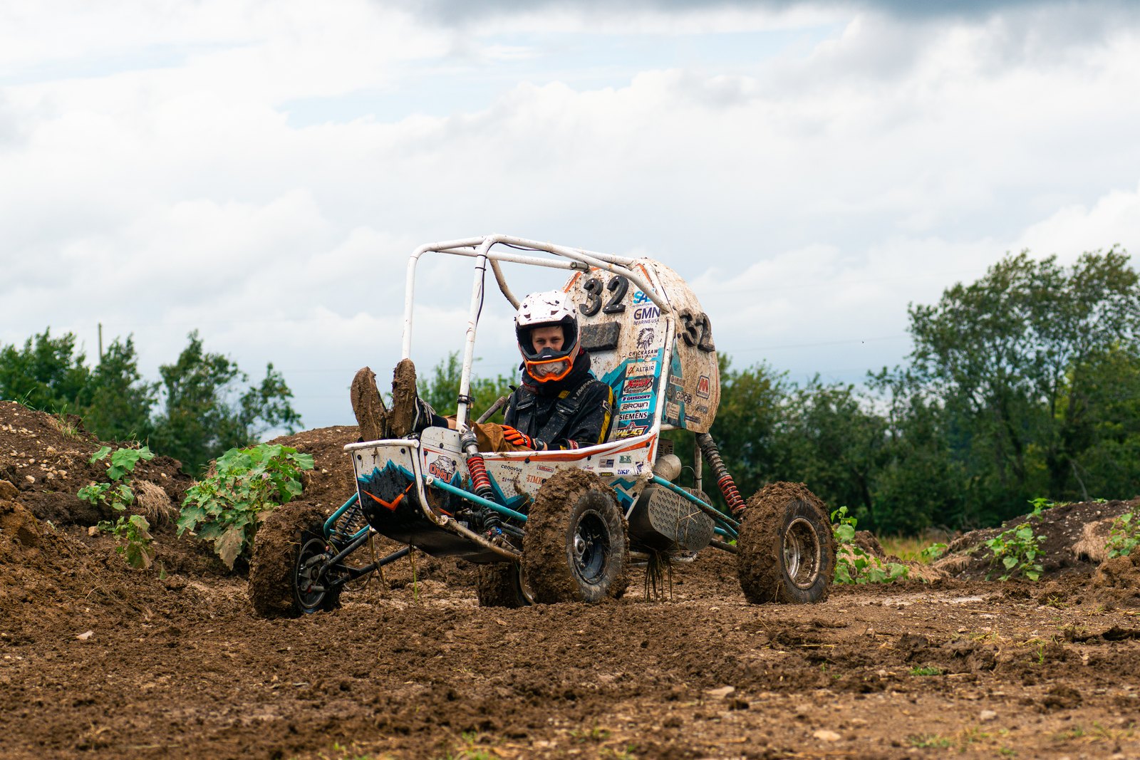 Baja car number 32 climbing a dirt mound with the inside front wheel lifted, trees and cloudy sky behind