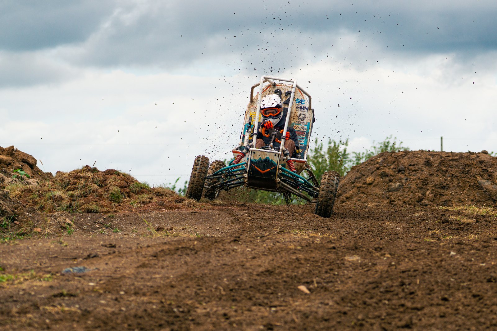 Baja car number 32 cresting a dirt mound head-on with one front wheel drooped and mud flying, under a cloudy sky