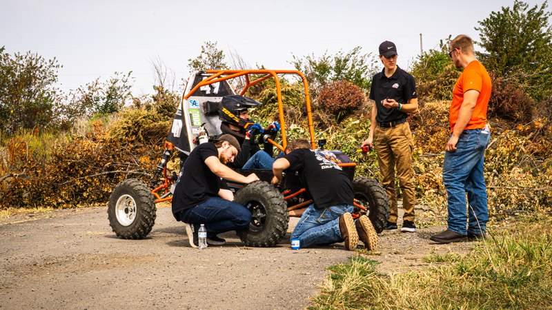Team members working on the orange Baja car beside a gravel road