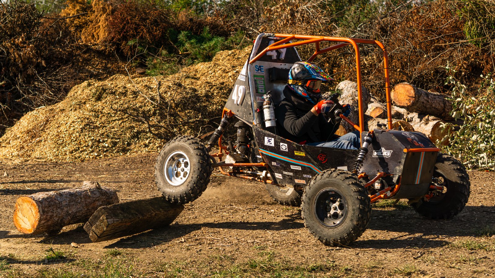 Baja car number 49 with its front wheel lifting over a log obstacle, showing suspension travel