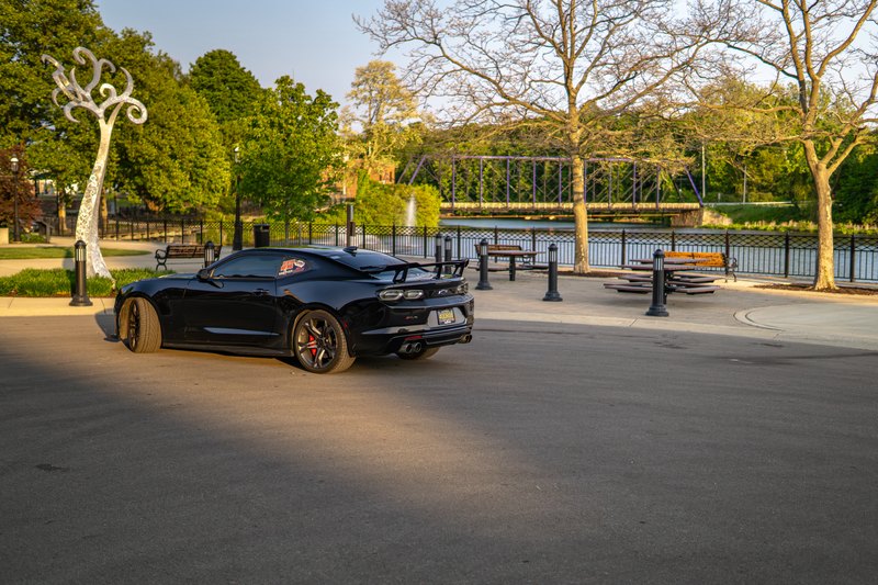 Black Camaro, rear three-quarter view showing the rear wing, in a park plaza with long shadows