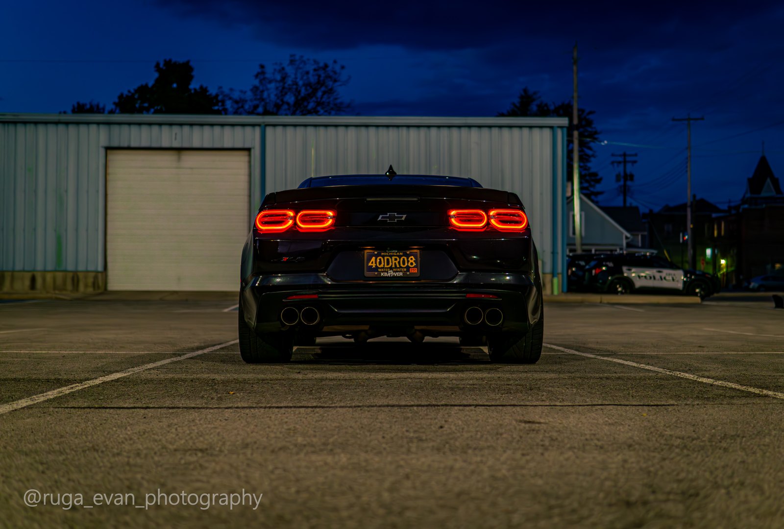 Rear of a black Camaro, taillights lit, centered in front of a garage door at dusk