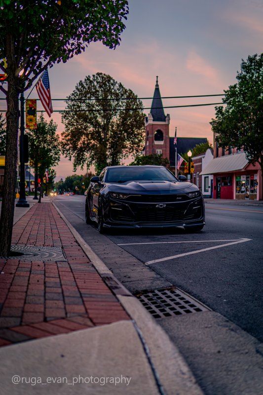 Brick sidewalk leading to a black Camaro with a church steeple and sunset behind