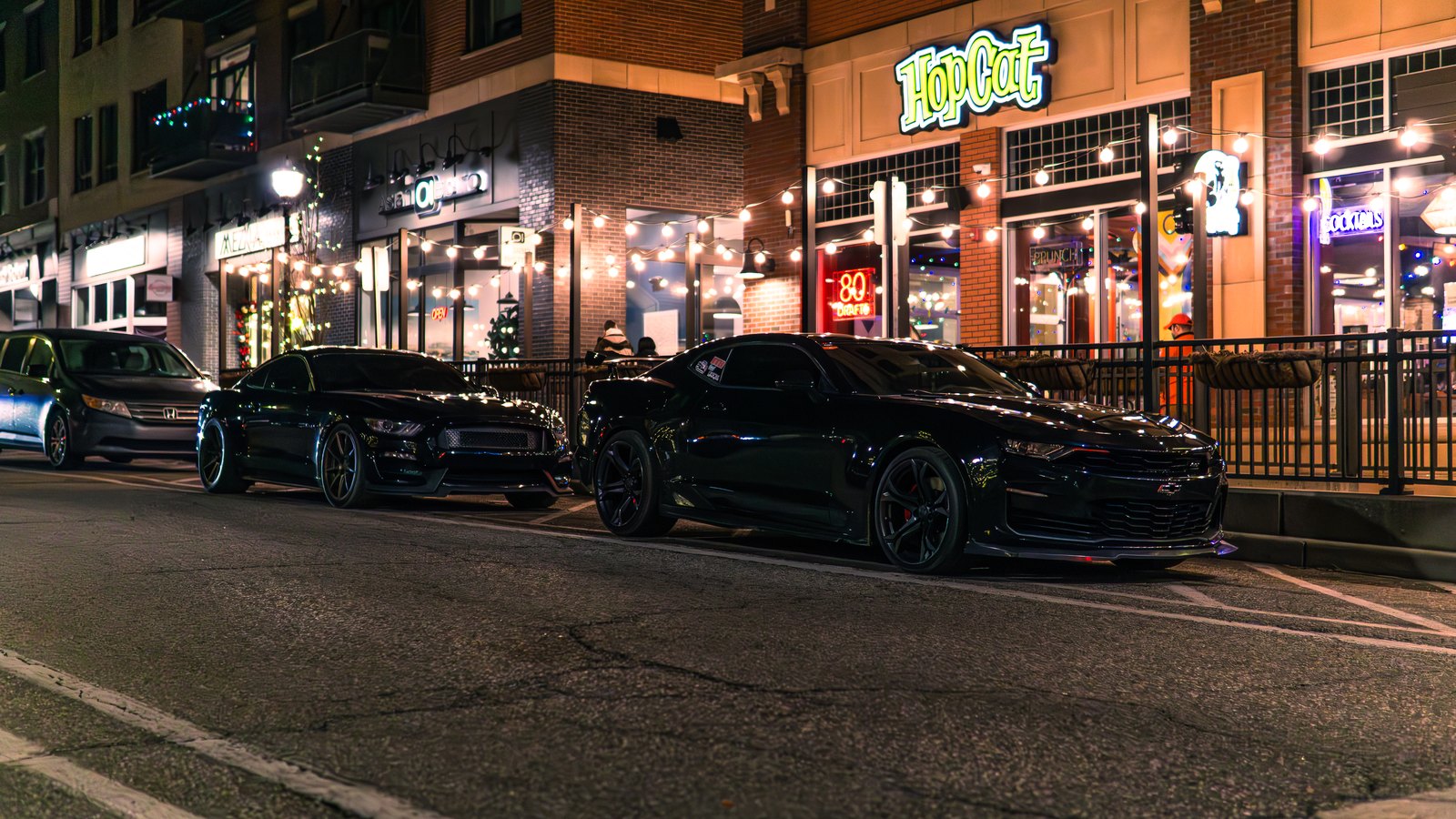 A black Mustang and a black Camaro parked together on a lit downtown street at night