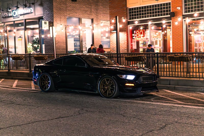 Low three-quarter view of a black Mustang under string lights in front of storefronts at night